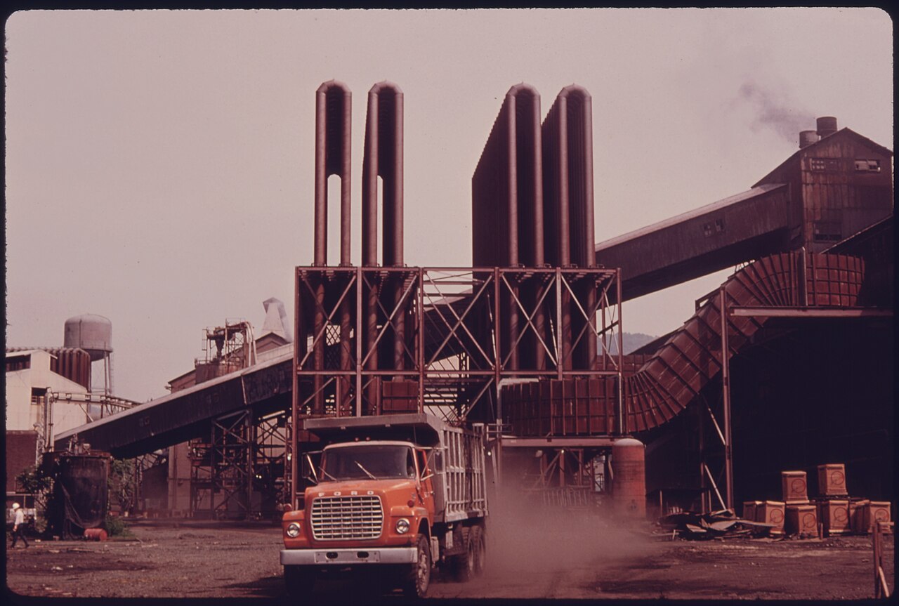A 1970s industrial plant photographed from the yard: conveyor galleries feeding a steel frame that carries four tall cylindrical emission stacks, with a dump truck raising dust in the foreground
