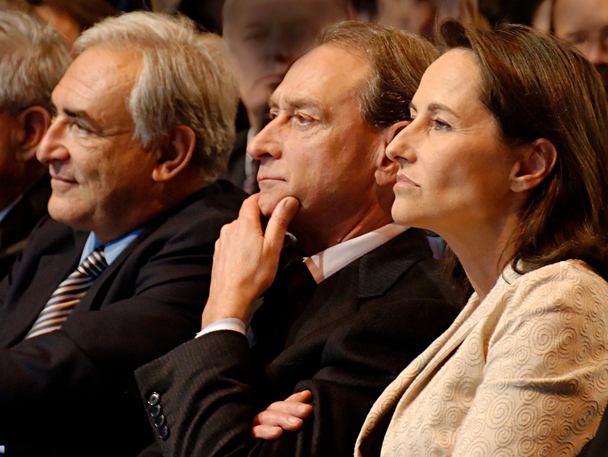 Three people seated side by side in the front row of a political meeting, listening intently, seen in close profile