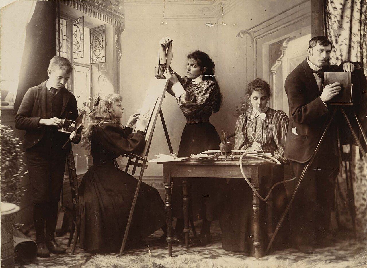 A posed nineteenth-century photograph of five people in a studio: one standing at a large plate camera, one at an easel, one holding up a print, one writing