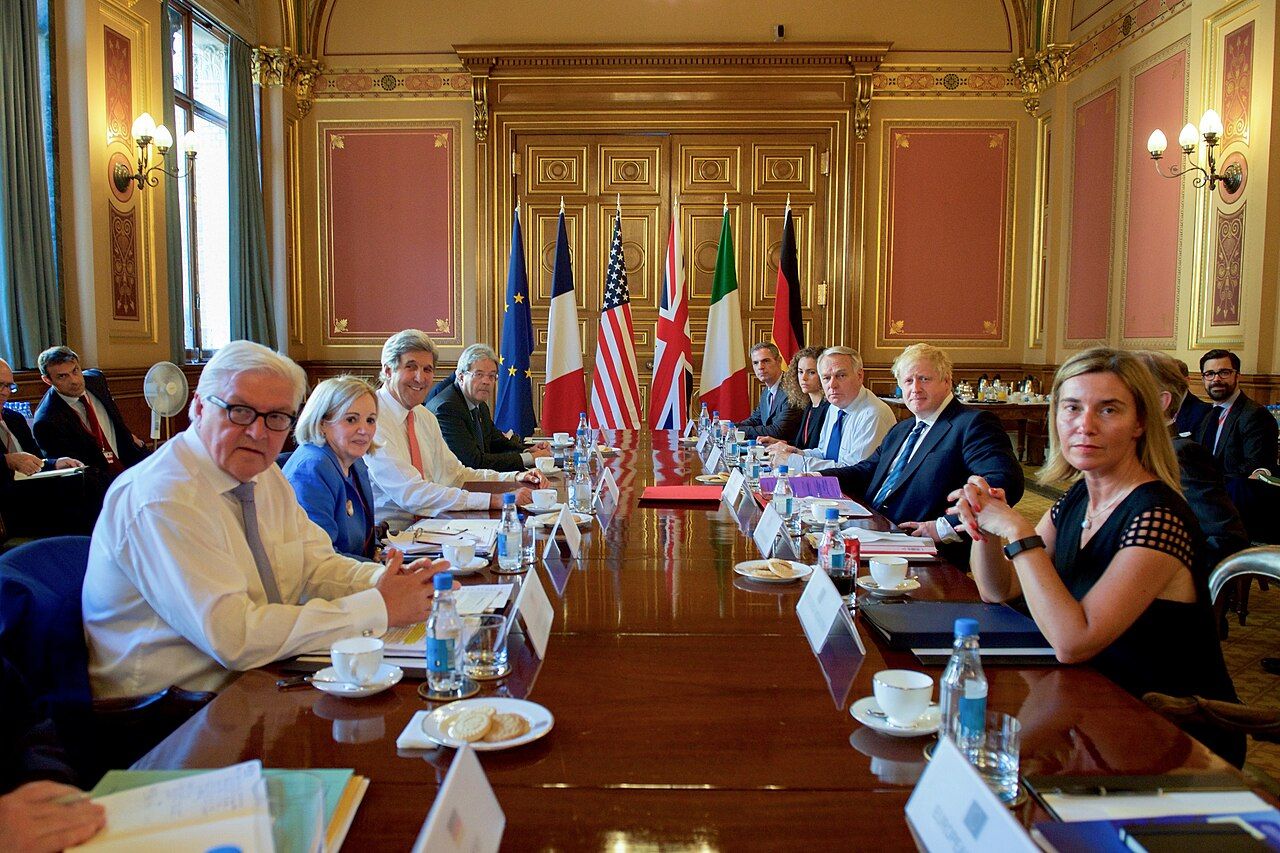 Foreign ministers and their delegations seated along both sides of a long conference table, with national flags behind them and briefing papers in front of each place.